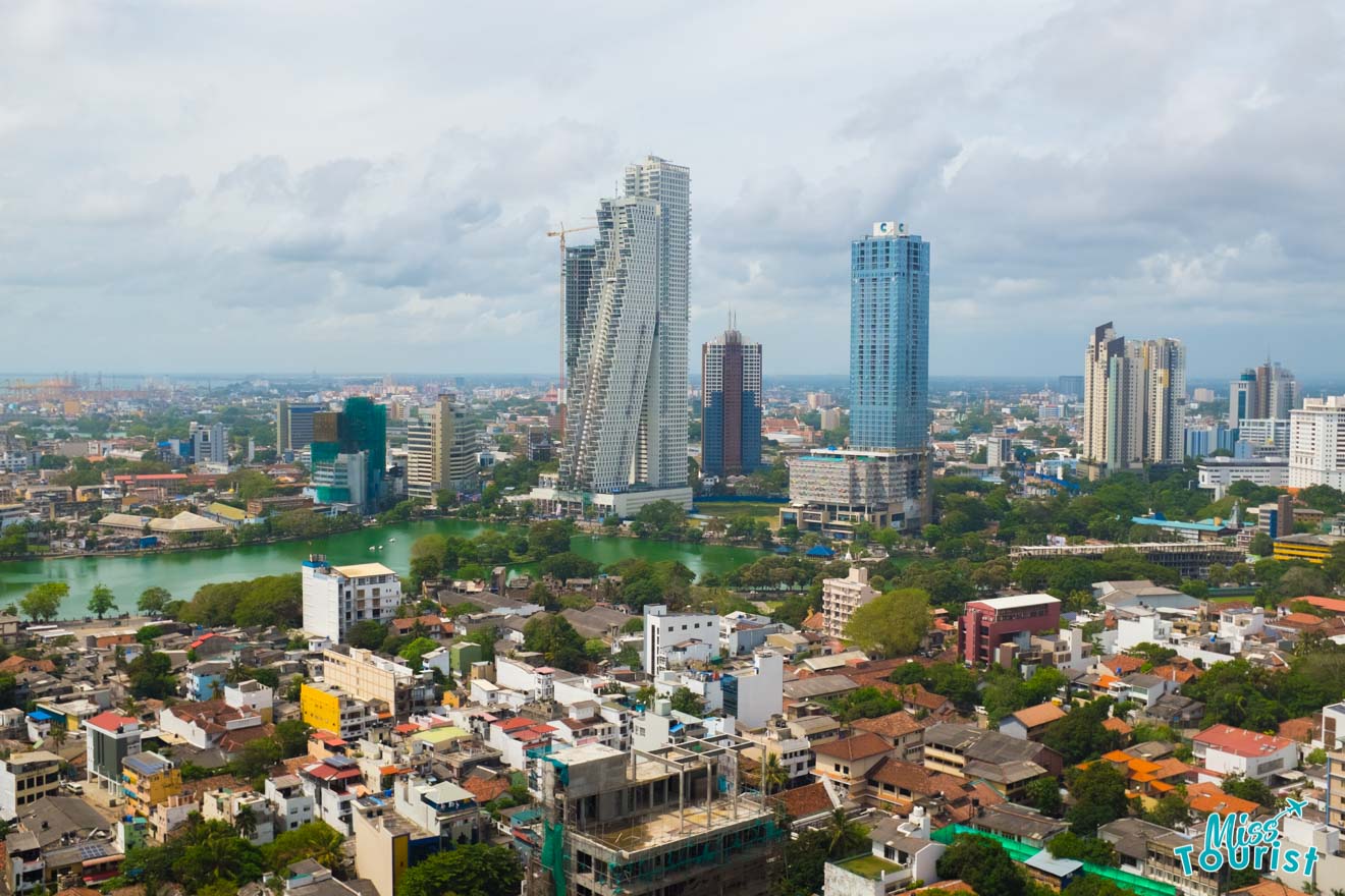 Alt text: "A panoramic view of Colombo's cityscape, featuring a mix of high-rise buildings, urban development, and areas of greenery, with a lake nestled among the city structures, under a partly cloudy sky.