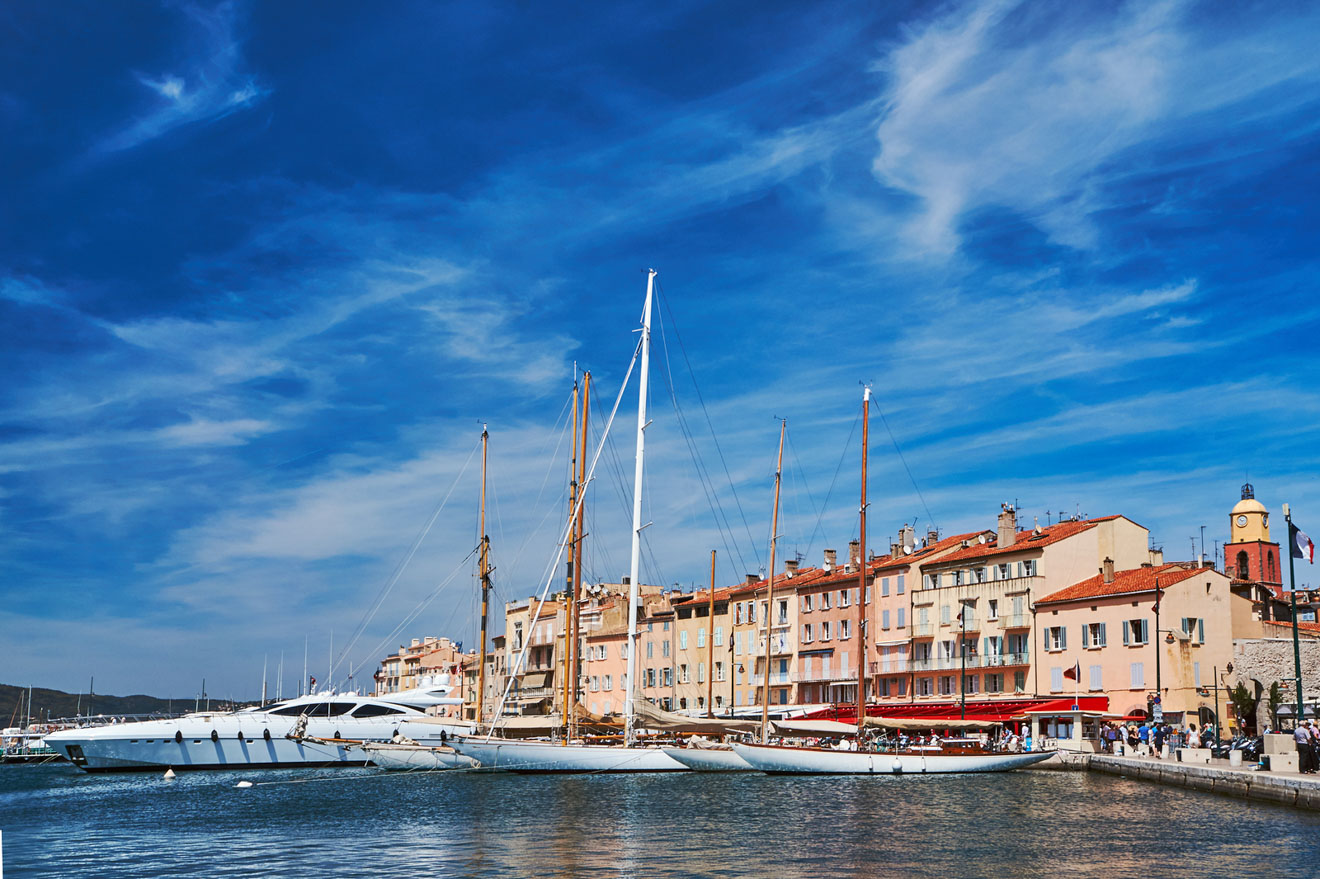 The Saint-Tropez harbor with luxury yachts moored in clear blue waters, backed by colorful traditional buildings under a bright blue sky with wispy clouds