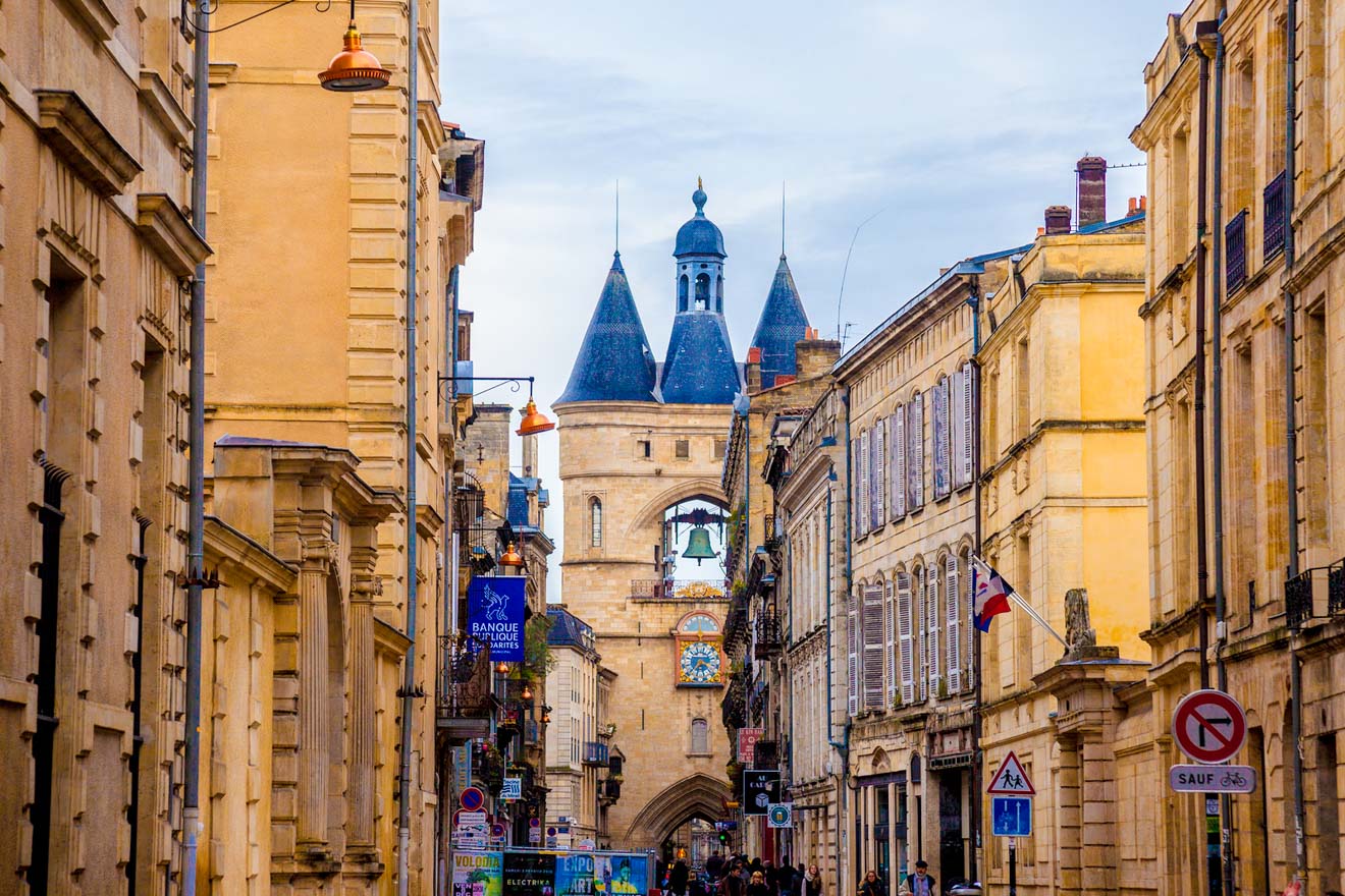 A narrow street in Bordeaux with a clock tower and a bell in the background.