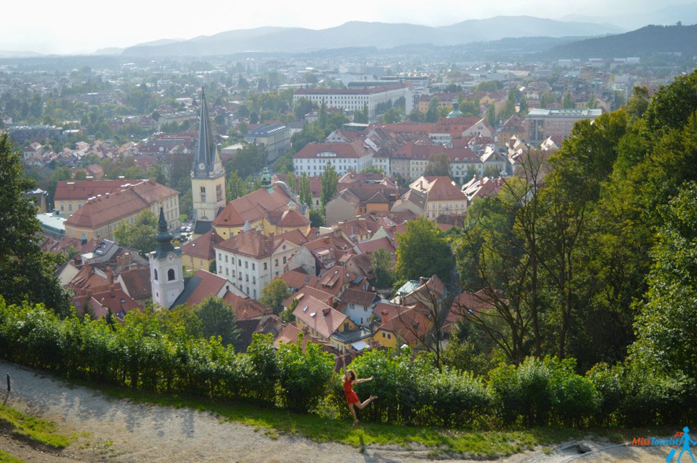 A person in a red shirt sits on a bench overlooking a city with a mix of historical and modern buildings, surrounded by trees and greenery, with mountains in the background.