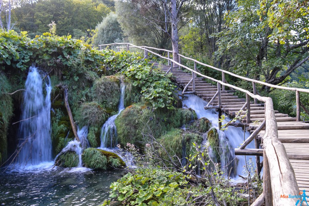 Wooden boardwalk meandering through dense greenery and multiple waterfalls at Plitvice Lakes
