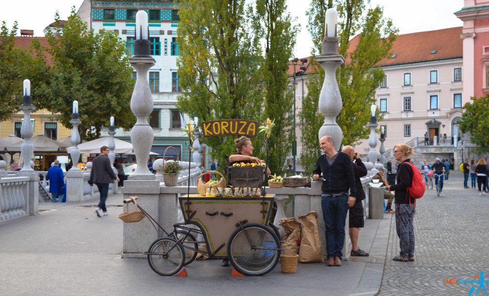 A street vendor sells corn from a decorated cart on a cobblestone square. Several people stand nearby, with trees and buildings in the background. A bicycle is attached to the cart.