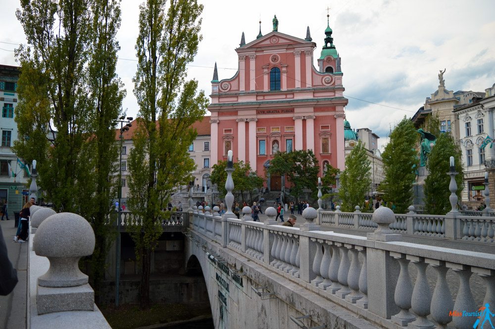 Baroque-style pink church building behind a stone pedestrian bridge lined with balustrades and trees on either side. People are walking on the bridge, and the sky is partly cloudy.