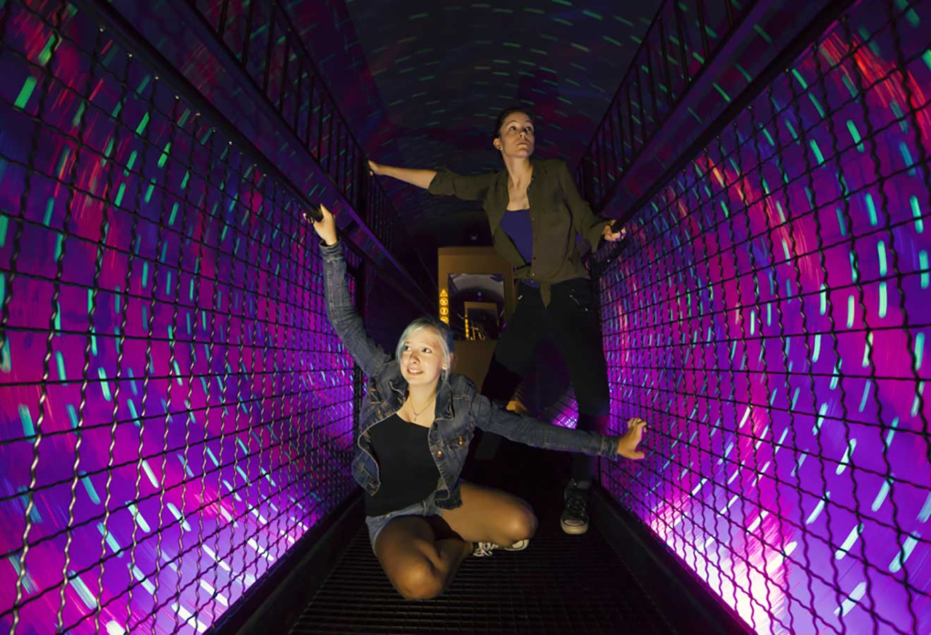 Two people pose energetically in a tunnel with vibrant purple and blue lights reflected on the walls. One stands with arms extended, while the other crouches with one arm raised.