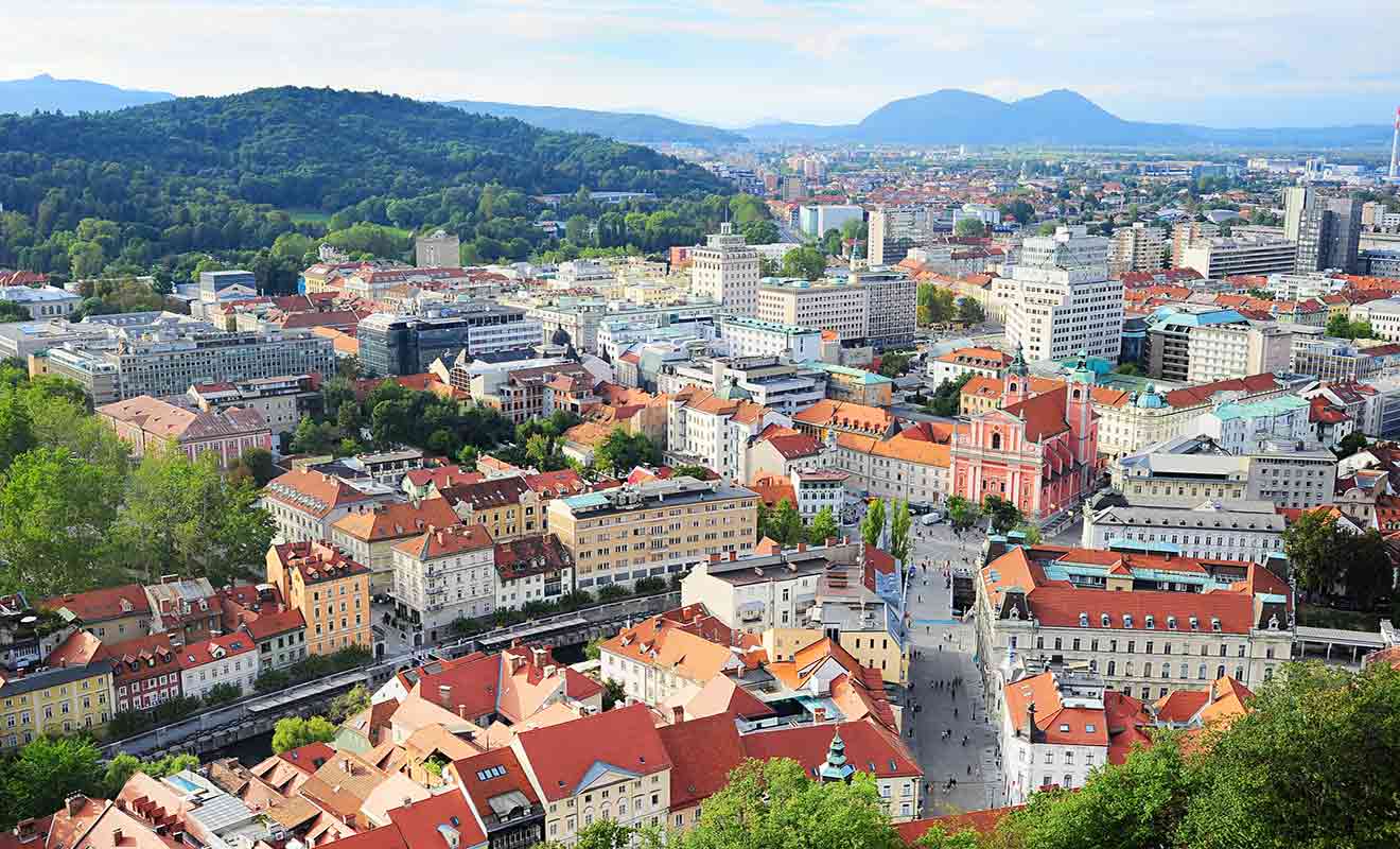 Aerial view of a city with a mix of modern and historic buildings, red-tiled roofs, a central square, and surrounding green hills.