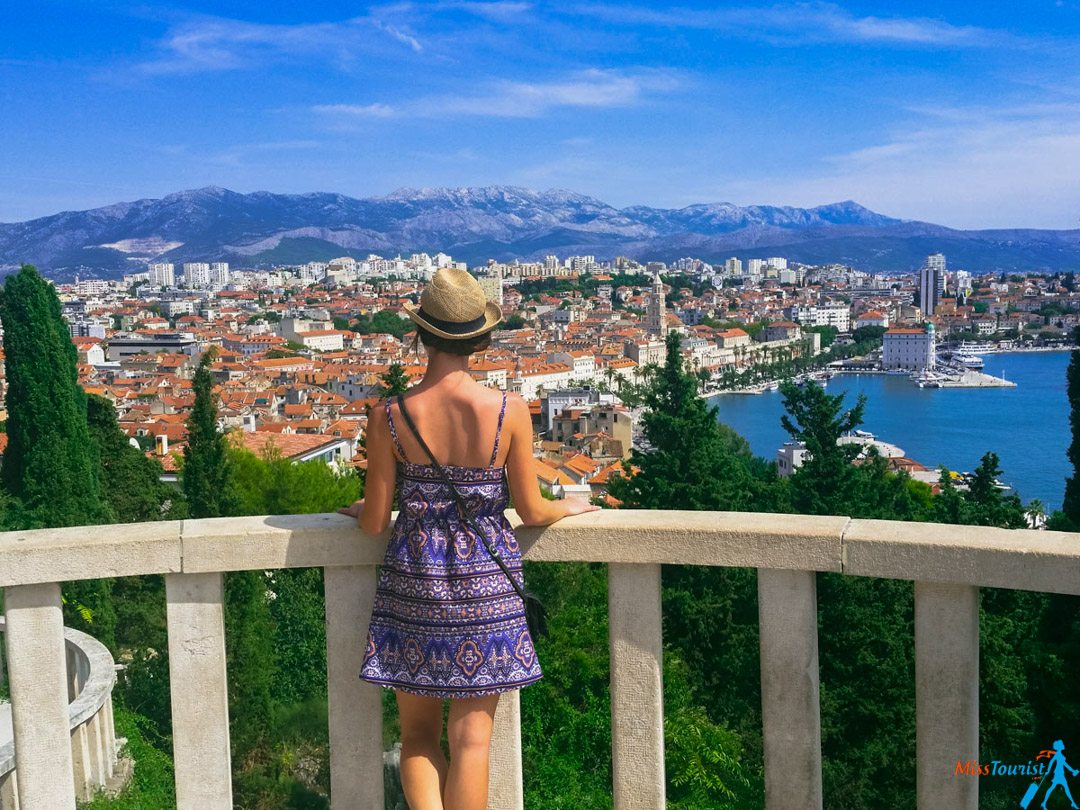 The author of the post in a patterned dress and hat stands on a balcony, overlooking a city with a coastal view and distant mountains.