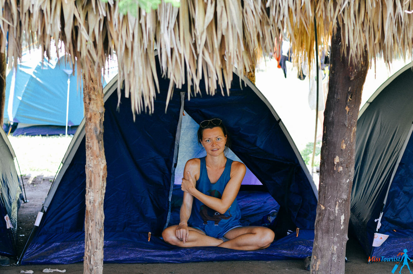 author of the posts sits cross-legged at the entrance of a blue tent under a thatched roof. She is wearing a blue top and sunglasses on her head, with other tents visible in the background.