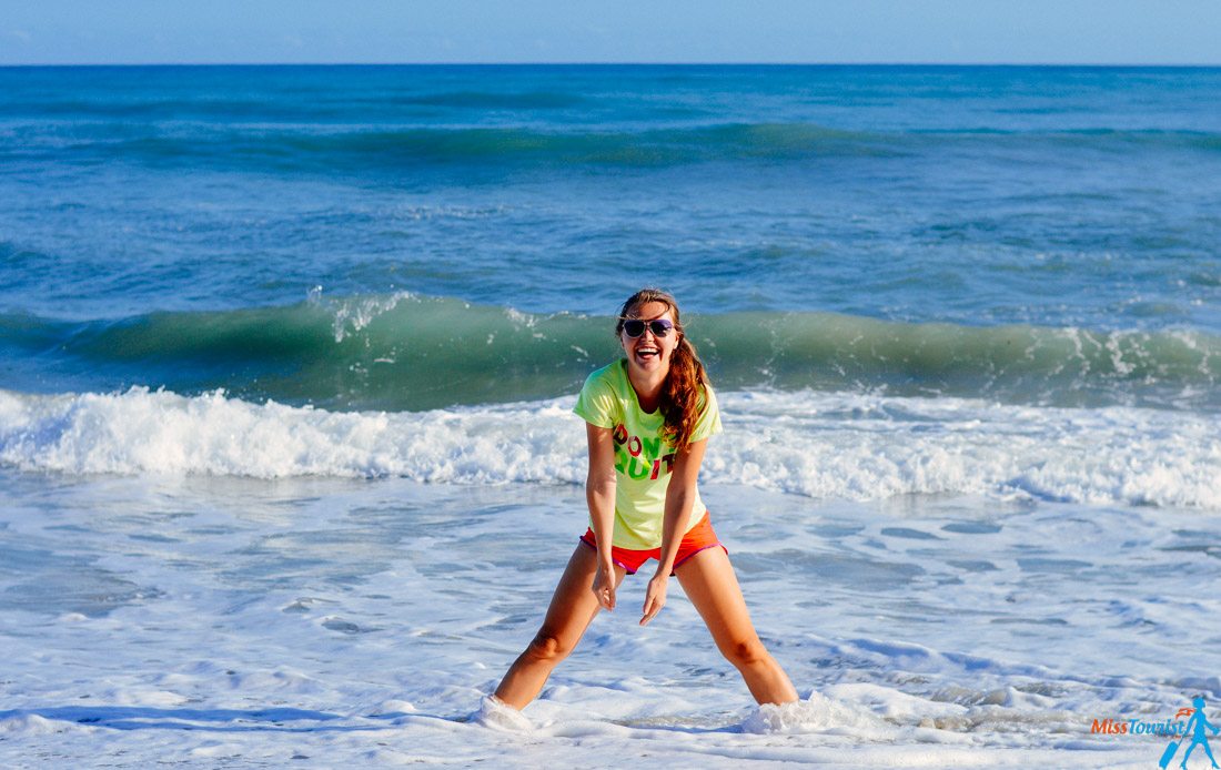 author of the posts wearing sunglasses, a green shirt, and red shorts stands smiling in shallow ocean water with waves in the background on a sunny day.