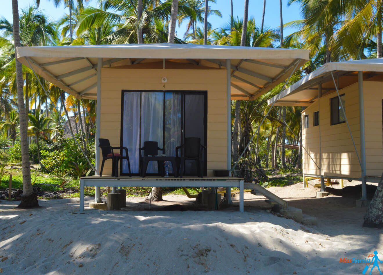 A small beach hut with a raised platform, a sliding glass door, two chairs and a small table on the porch, surrounded by palm trees and sand in a tropical setting.