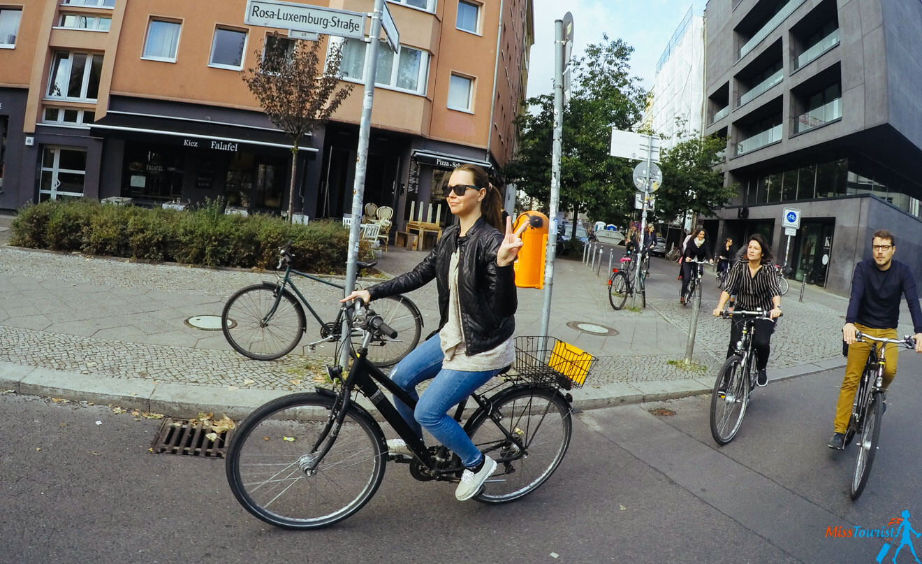 The founder of the website, Yulia, rides bicycles on a city street, with buildings and street signs in the background. The leading cyclist, wearing a leather jacket and sunglasses, gestures with a peace sign.