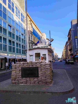 Checkpoint Charlie in Berlin, with a soldier mannequin and sandbags in front of a historical guardhouse. Surrounding buildings are visible under a clear sky.