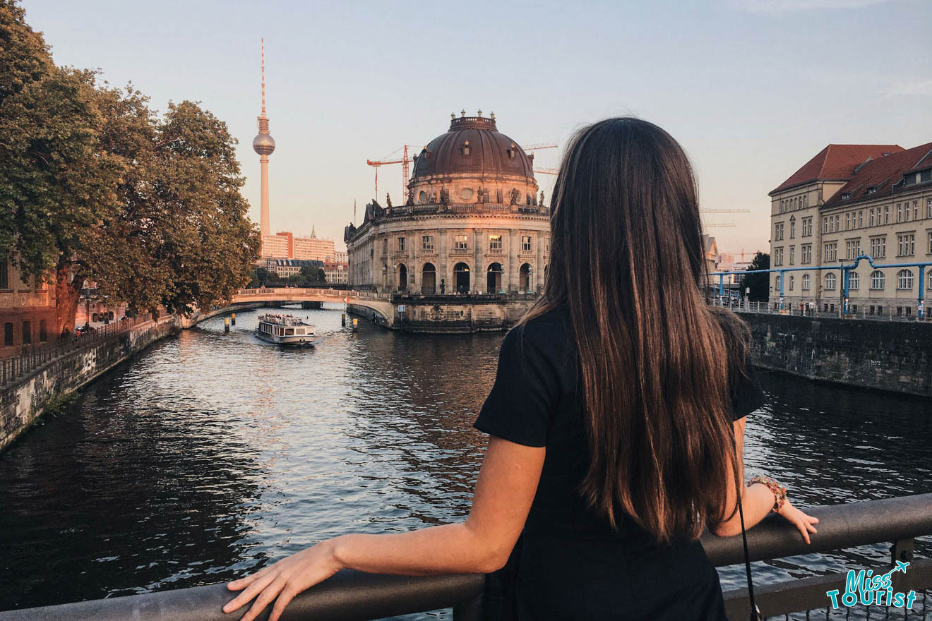 A person stands on a bridge overlooking a river with a boat, historic building, and TV tower in the background.