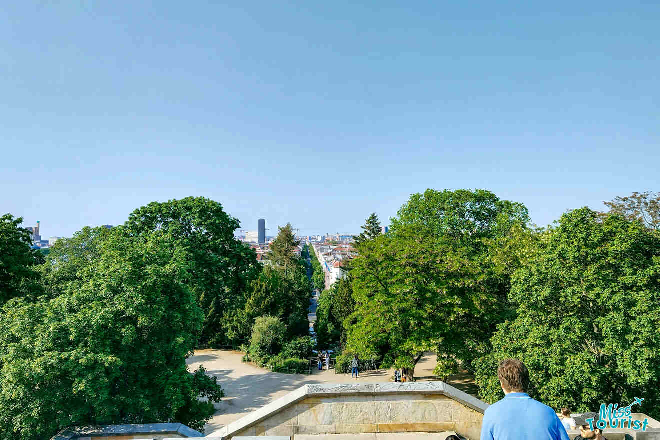 A person stands on a terrace overlooking a tree-filled park with a cityscape in the distance under a clear blue sky.