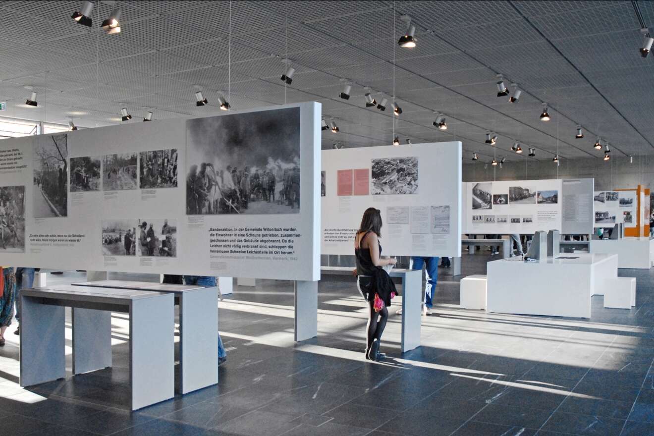 People view large informational displays and historical photos in a modern, spacious museum exhibit with natural light and hanging ceiling lights.