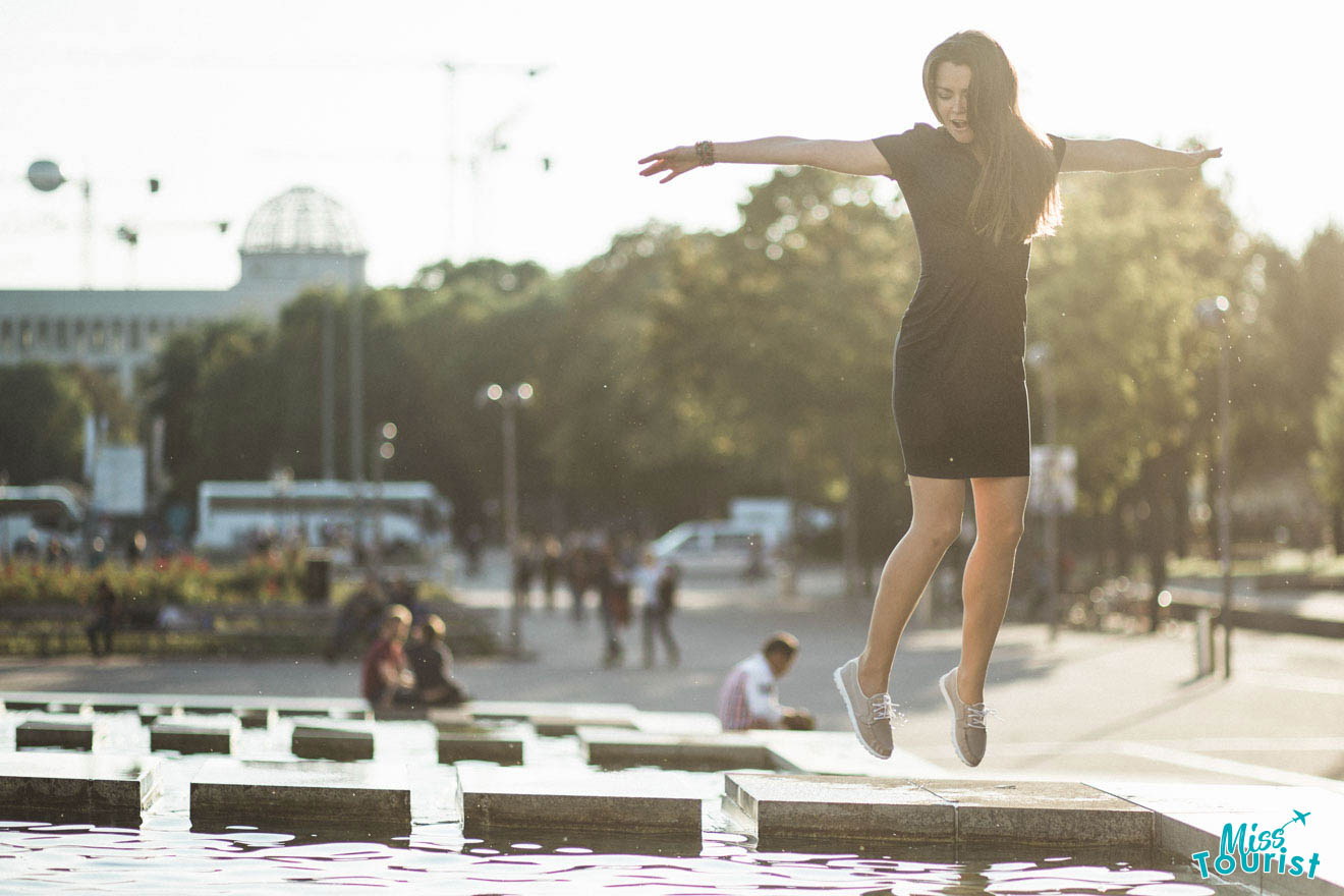 A woman in a black dress appears to be leaping or balancing on the edge of a fountain in a public park. People and trees are visible in the background.