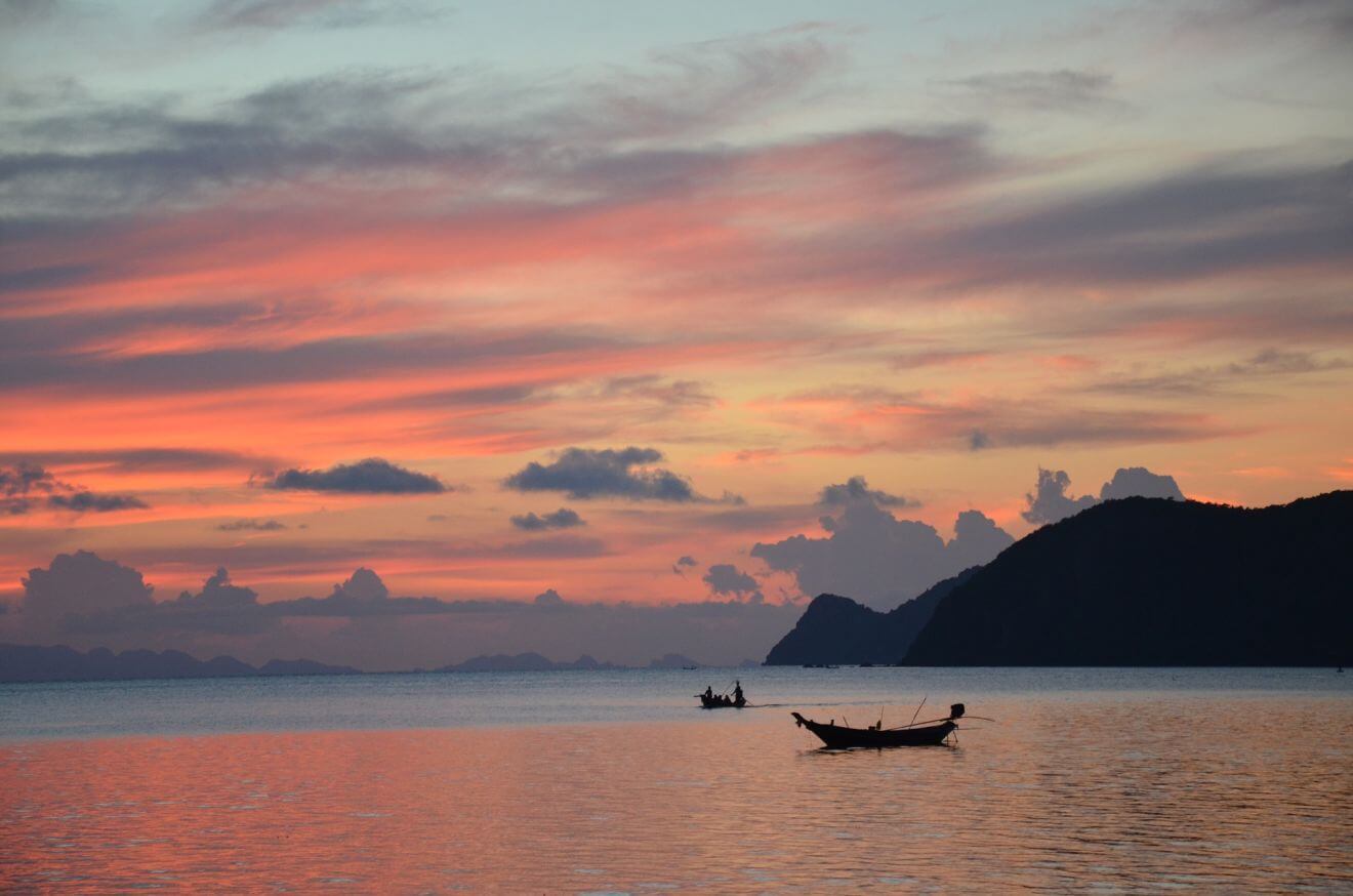 A serene sunset in Koh Phangan with a vibrant display of pink and orange hues reflecting over calm waters, with silhouettes of distant islands and a traditional long-tail boat in the foreground