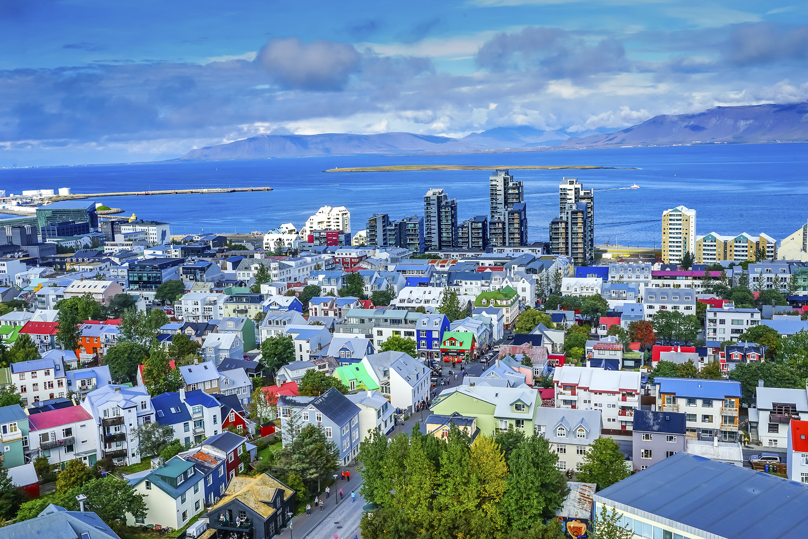 A vibrant city panorama showcasing an array of colorful buildings with a backdrop of blue sea and mountains under a partly cloudy sky