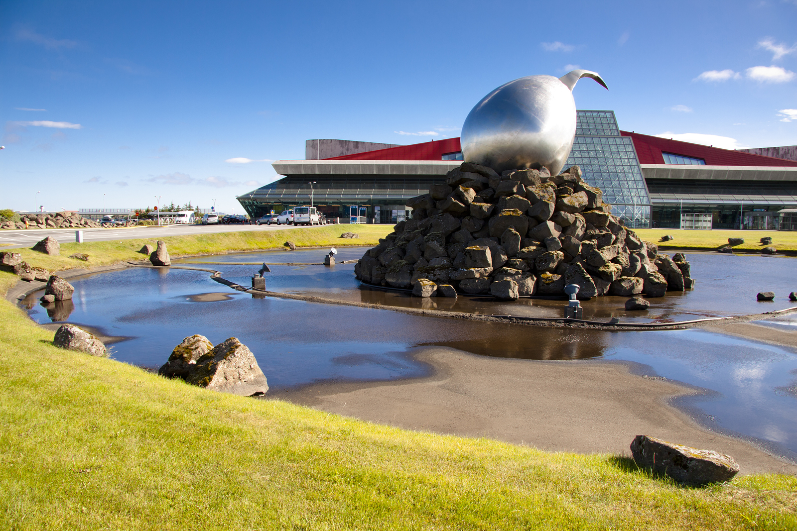 A serene outdoor sculpture of a bird perched on a sphere, set amidst a reflective pool with rocks, with an airport building in the background under a clear blue sky