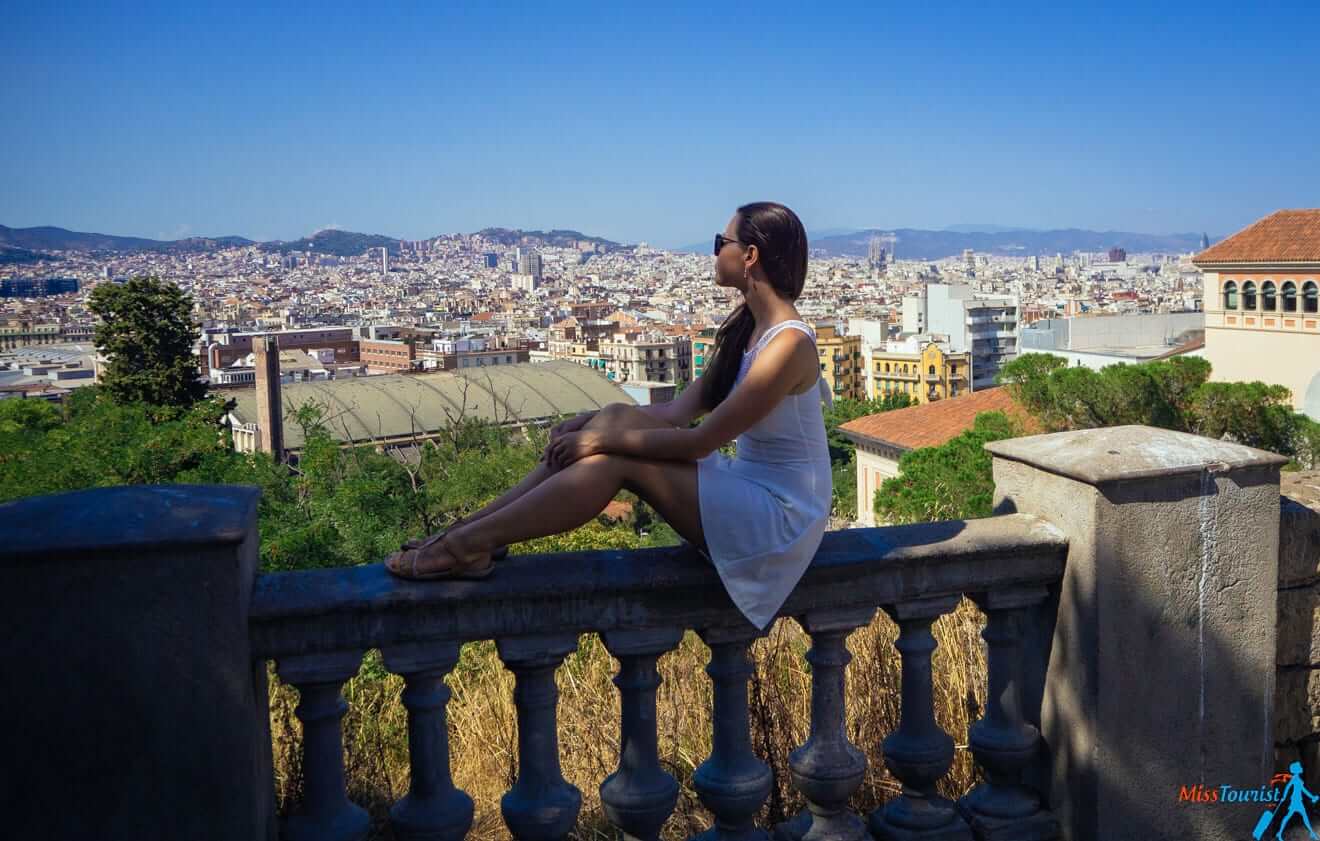 author of the post in a white dress sits on a stone railing overlooking a cityscape on a clear day.