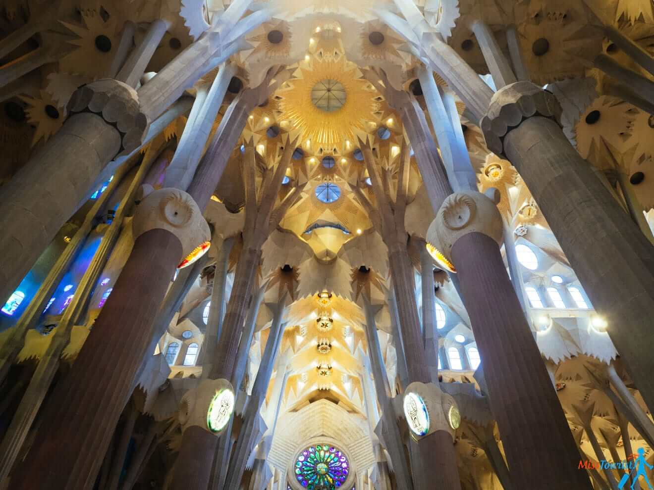 Interior of a grand cathedral with detailed architectural design, tall columns, arched ceilings, stained glass windows, and intricate patterns illuminated by natural light.