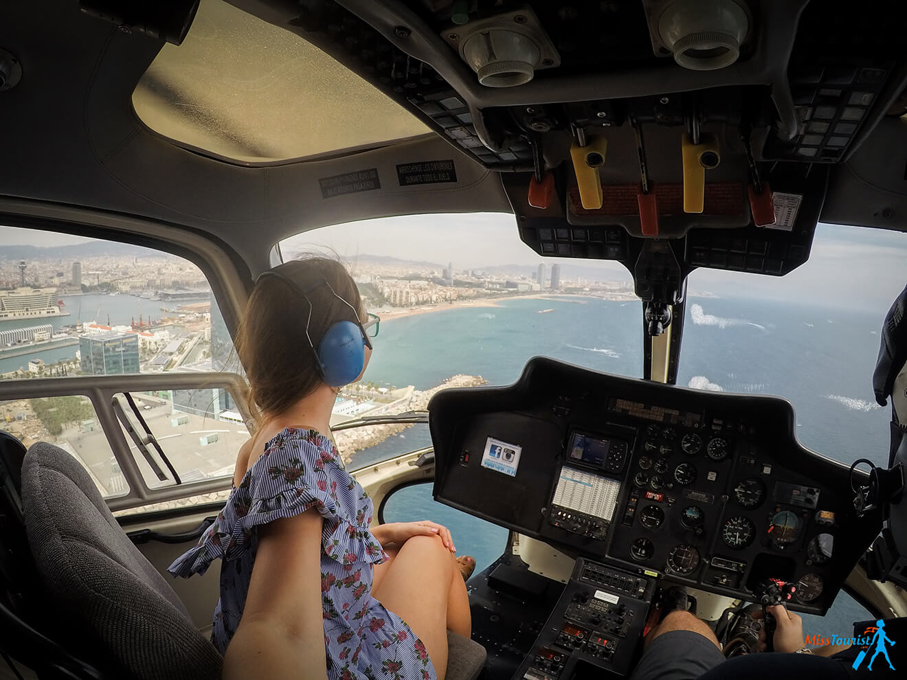 author of the post wearing headphones is seated in the front of a helicopter, looking out at a coastal city and ocean below. The pilot is visible operating the controls.