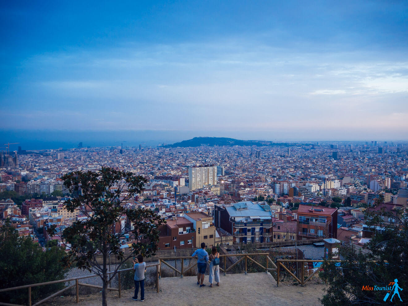 Three people stand at an overlook, observing a vast cityscape at dusk, with buildings stretching to the horizon under a partly cloudy sky.