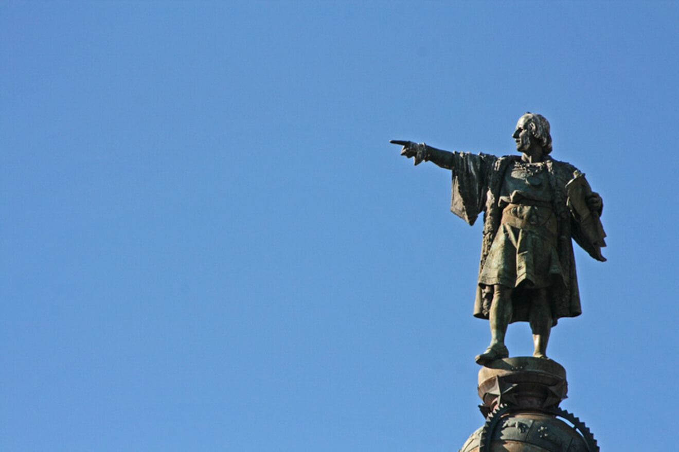 A bronze statue of a historical figure dressed in period clothing stands on a pedestal, pointing into the distance against a clear blue sky.