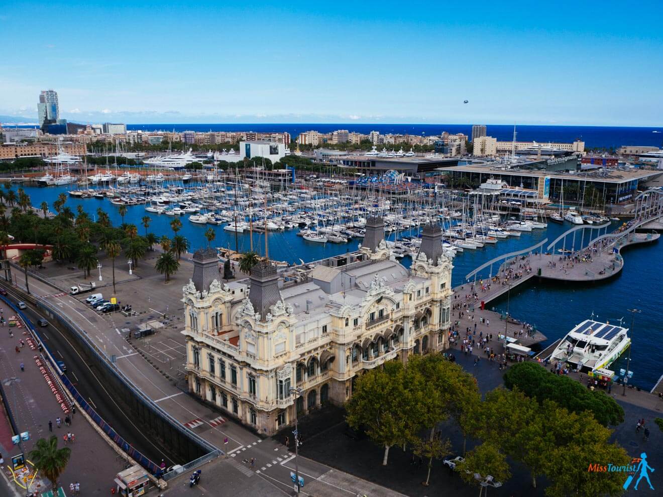 Aerial view of a marina with numerous docked boats, a large historic building in the foreground, and a cityscape with low-rise buildings in the background on a clear day.