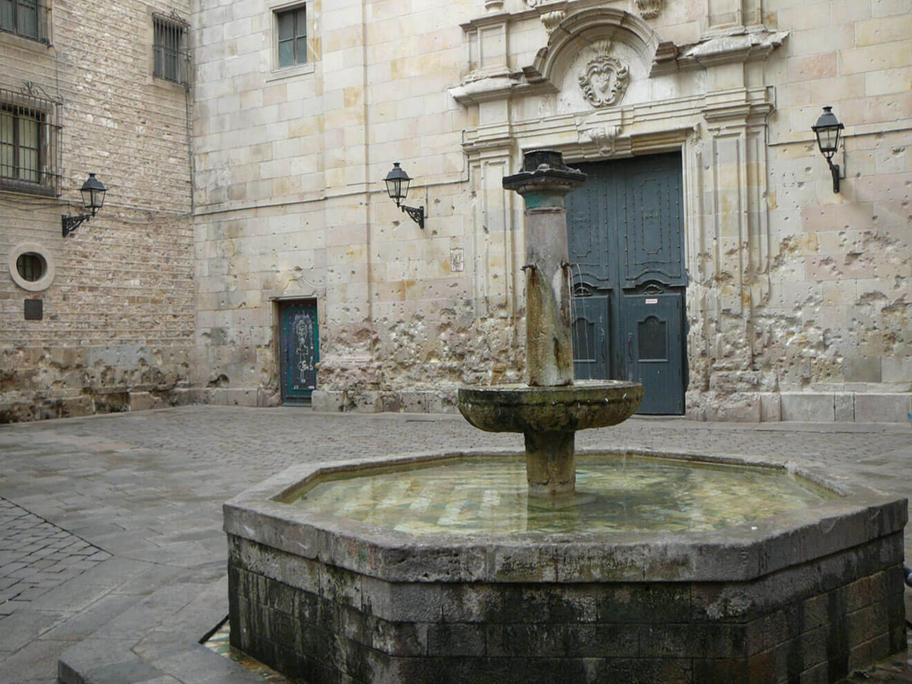 A stone fountain stands in the center of a cobblestone courtyard with worn beige brick walls and two closed doors in the background.