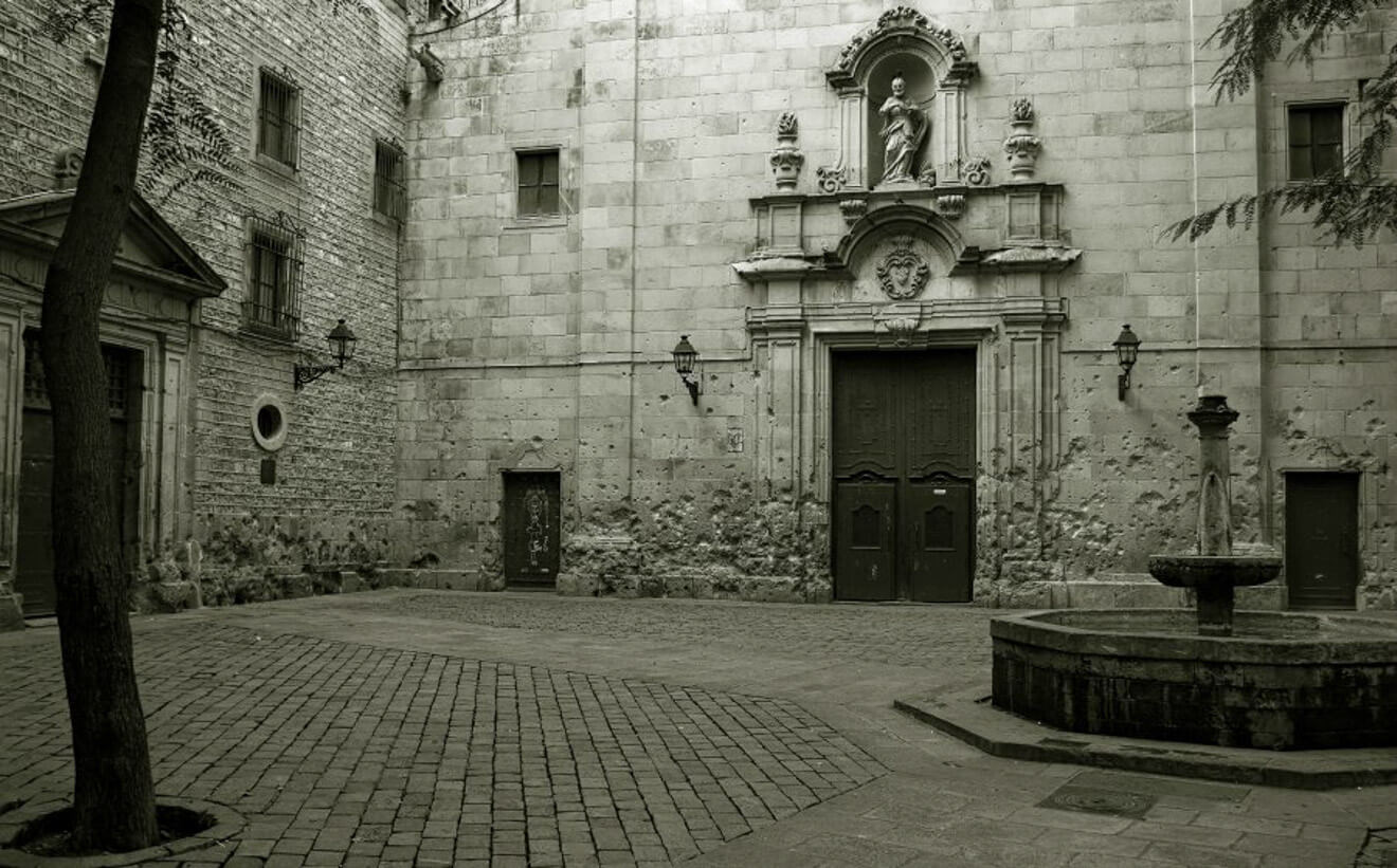 A stone courtyard with a weathered building, featuring a statue above a large wooden door. The area includes a cobblestone ground, a tree on the left, and a round stone fountain on the right.