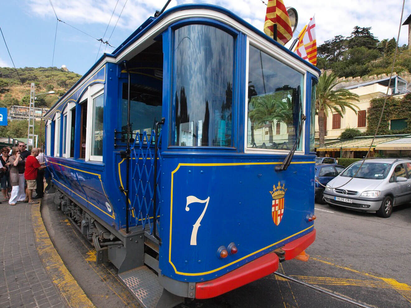 Blue and white tram with the number 7 on the side, parked at a station. Flags fly above, and people, cars, and buildings are visible in the background.