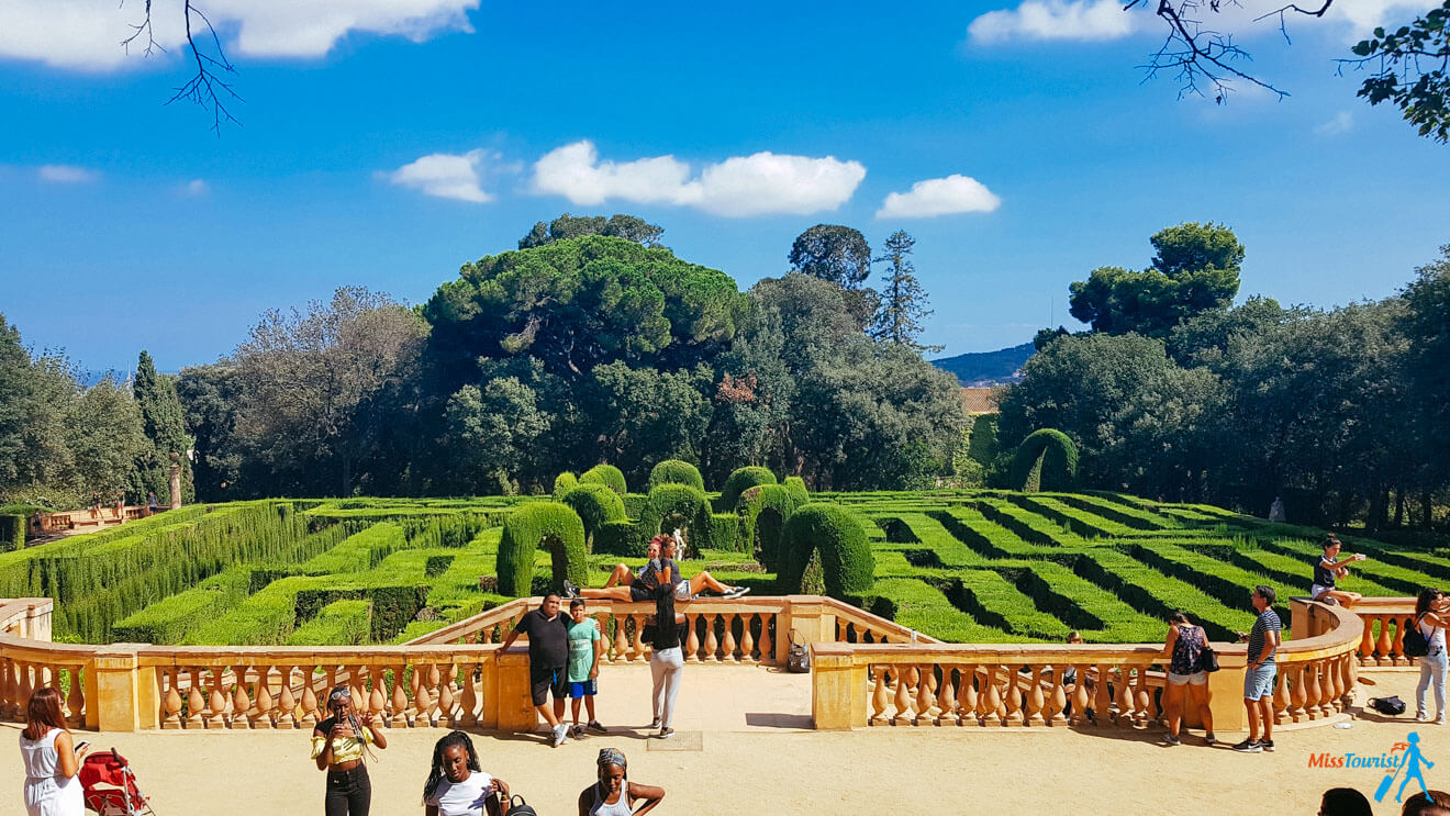 A group of people stand and walk around a hedge maze in a park, surrounded by lush greenery and blue sky with scattered clouds. Some are taking photos while others are enjoying the view.