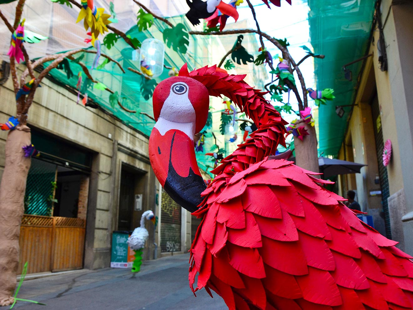 Vibrant red parrot sculpture with detailed feathers on a decorated street with colorful bird cutouts and green net covering.