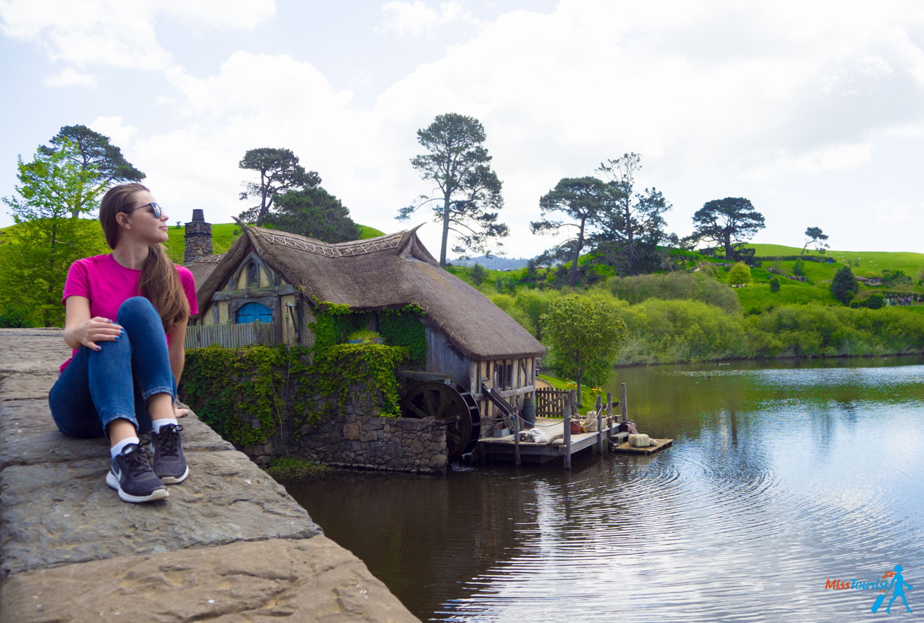 a woman sitting on a bridge near the water and the hobbit house in the background