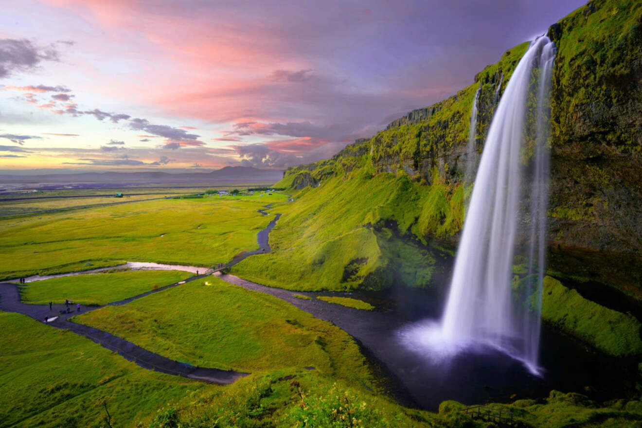 A majestic waterfall cascades down a lush, moss-covered cliff into a serene pool below, with a vivid sunset illuminating the expansive green landscape in Iceland