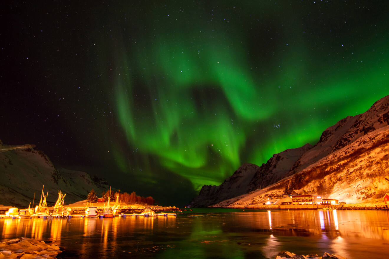 The Northern Lights (Aurora Borealis) dance in the night sky, casting a green glow over a snowy fjord with illuminated buildings in an Icelandic village