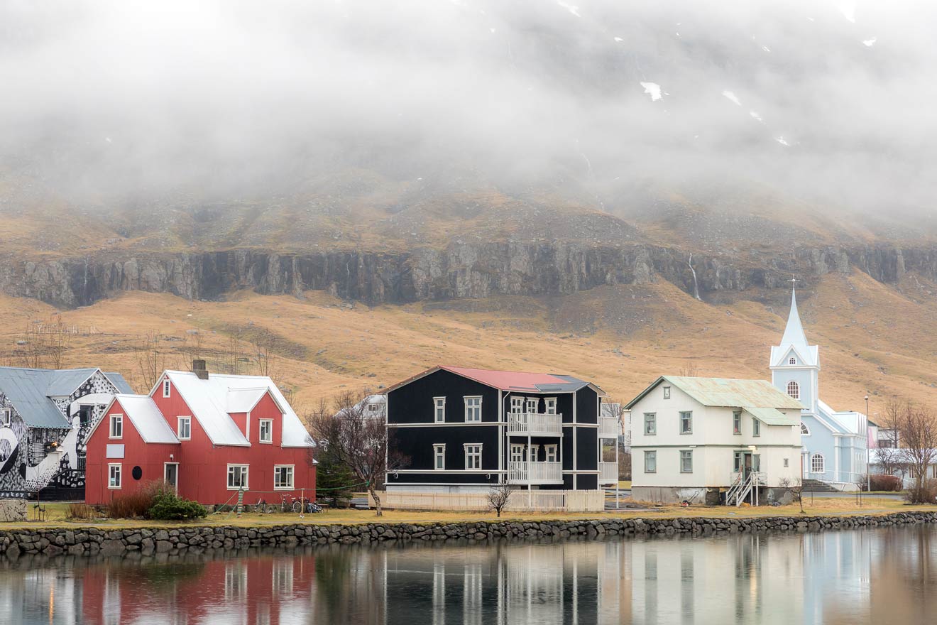 Charming houses in red, black, and white, reflecting on the still waters of a lake with misty, rugged mountains in the background, in a peaceful Icelandic village