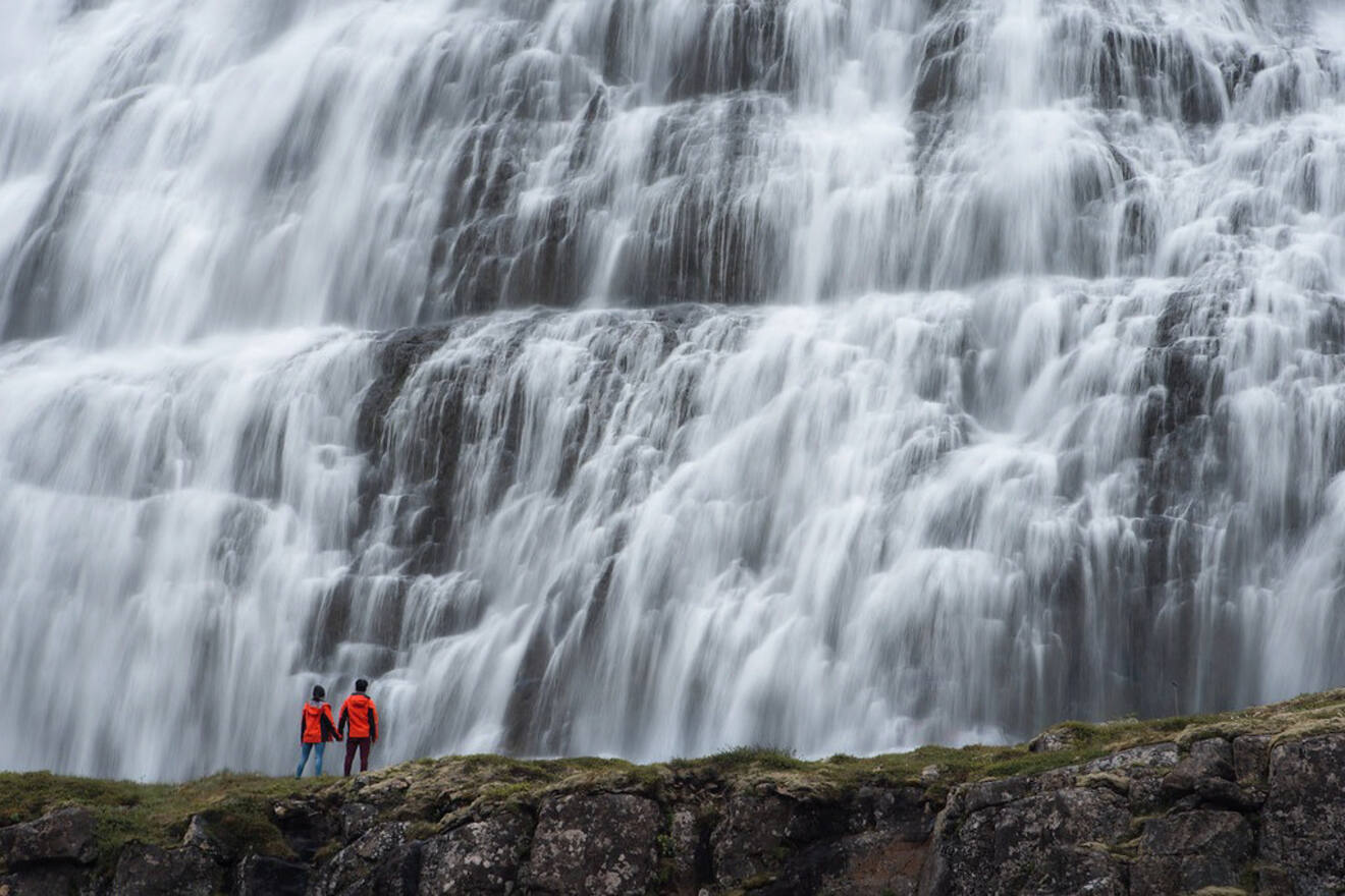 Two hikers in bright orange jackets stand in awe before the sprawling, textured cascade of Dynjandi waterfall in the Westfjords of Iceland