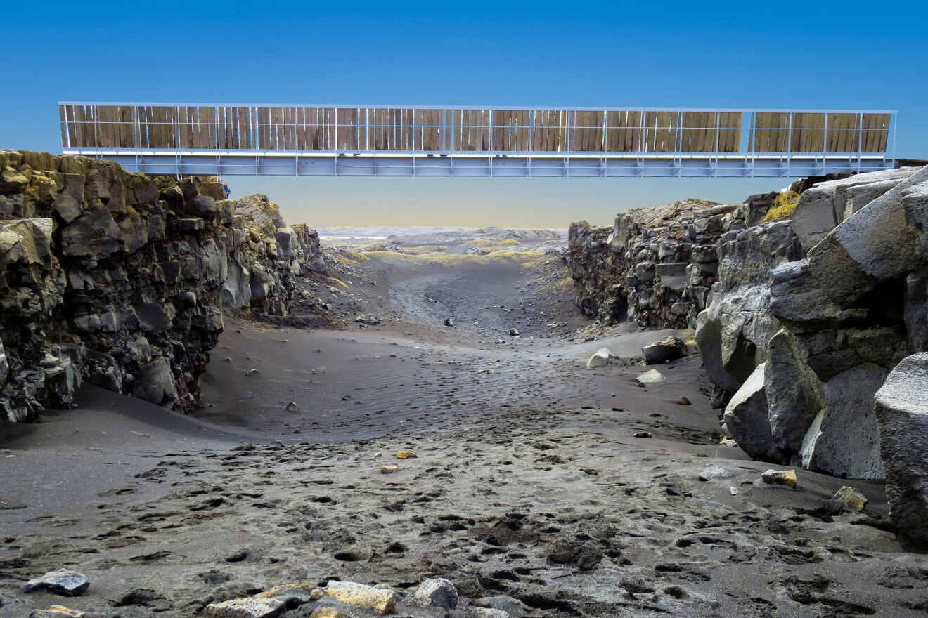 A modern pedestrian bridge spans a stark, dramatic rift between two tectonic plates with a barren, volcanic landscape under a blue sky in Þingvellir National Park, Iceland