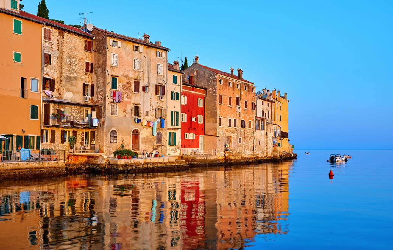 Historic buildings with weathered facades and colorful shutters reflect on the calm water at sunset in Rovinj, creating a tranquil and picturesque scene