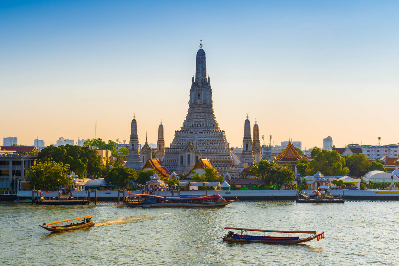 The Wat Arun temple in Bangkok, Thailand, at sunset with golden light illuminating its intricate spires and statues, with boats cruising on the Chao Phraya River.