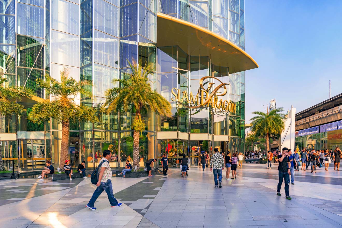 Exterior of Siam Paragon shopping mall in Bangkok with glass facade reflecting the sky, surrounded by palm trees and bustling with shoppers.