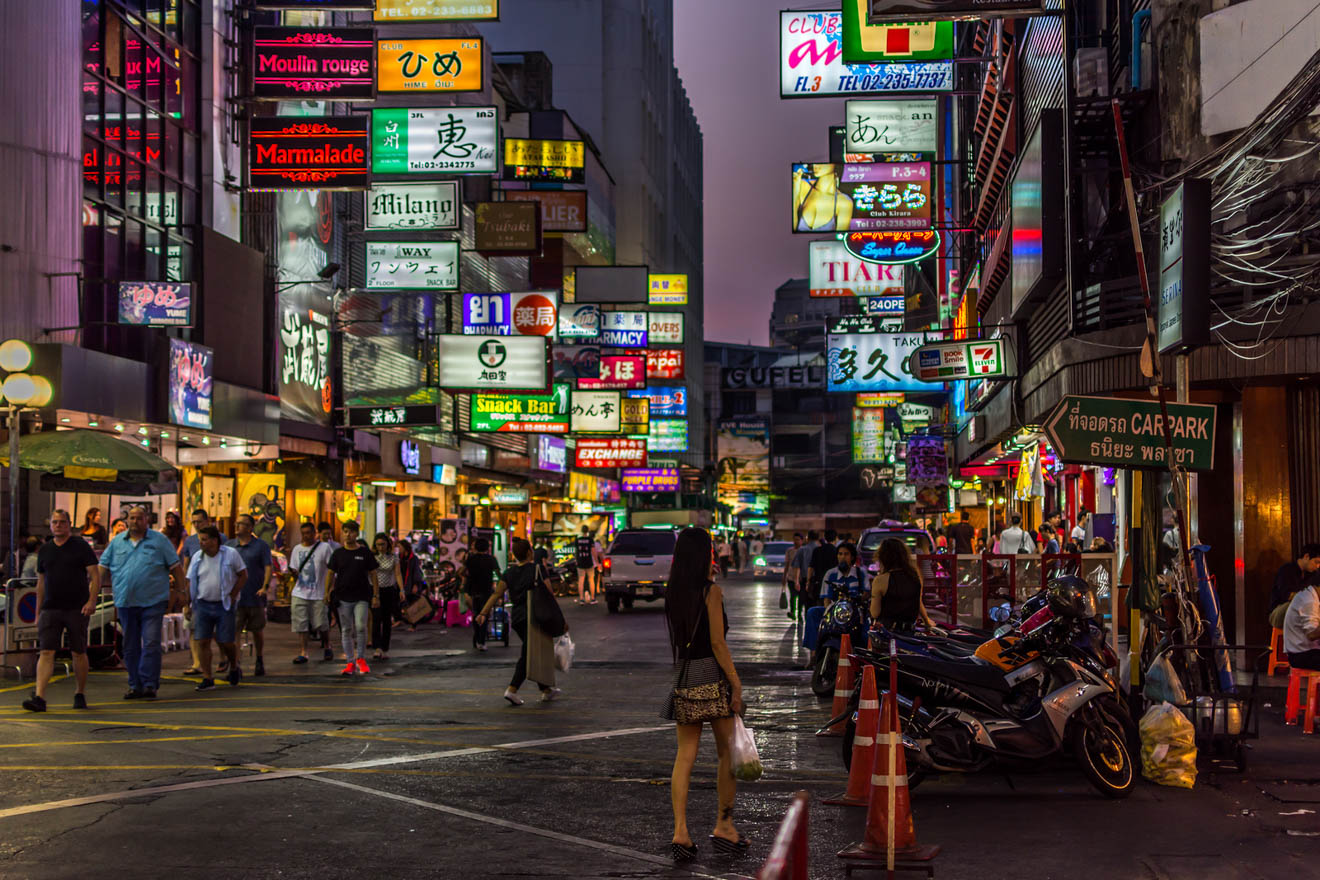 Neon-lit nightlife of Bangkok with vibrant signs in various languages, crowds of people, and a bustling street atmosphere.