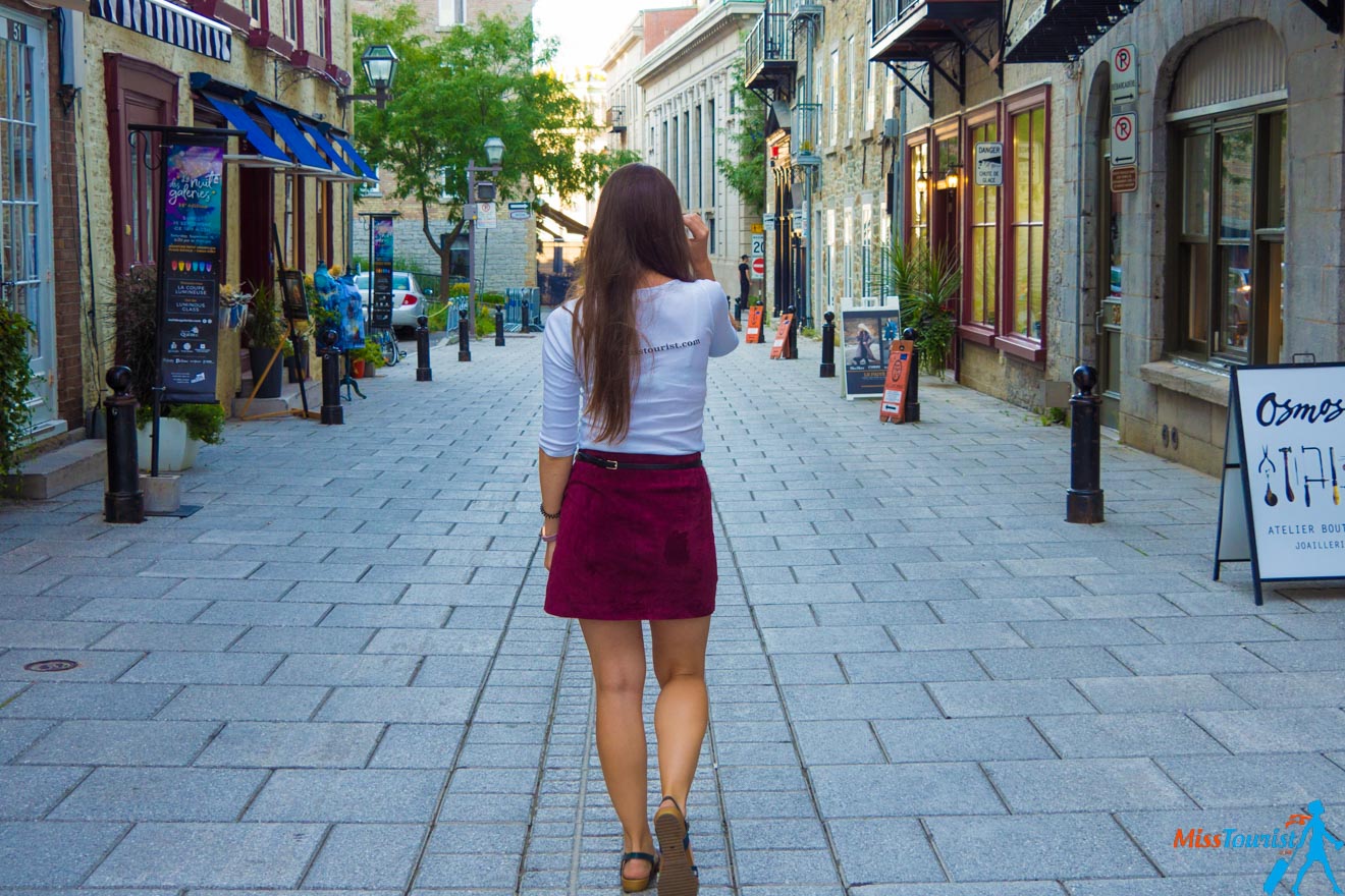 The writer of the post in a white top and burgundy skirt walking away down a charming cobblestone street lined with historic buildings and boutique shops.