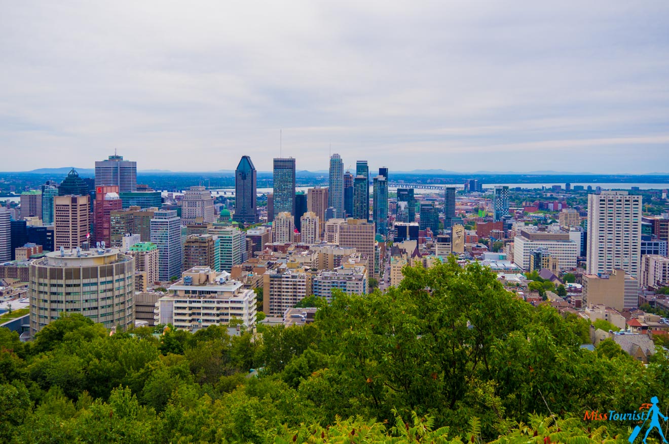 A panoramic cityscape view of Montreal from an elevated vantage point, showcasing a tapestry of buildings against a backdrop of distant mountains and overcast skies.