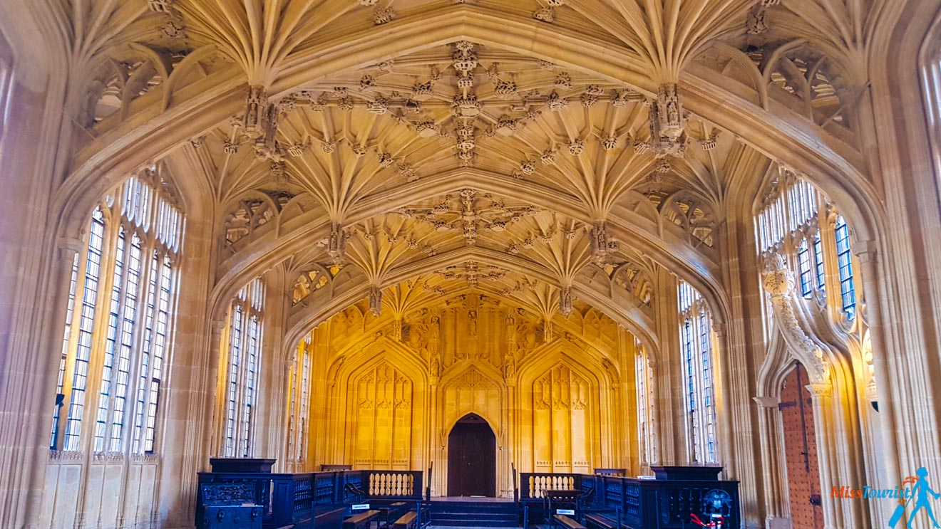 A large, ornate Gothic hall with a vaulted ceiling, intricate stone carvings, and tall, narrow arched windows. The room features wooden benches and a central doorway.