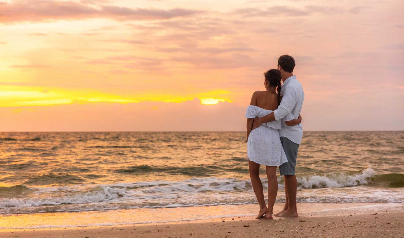 A couple hugging on the beach looking towards the ocean at sunset