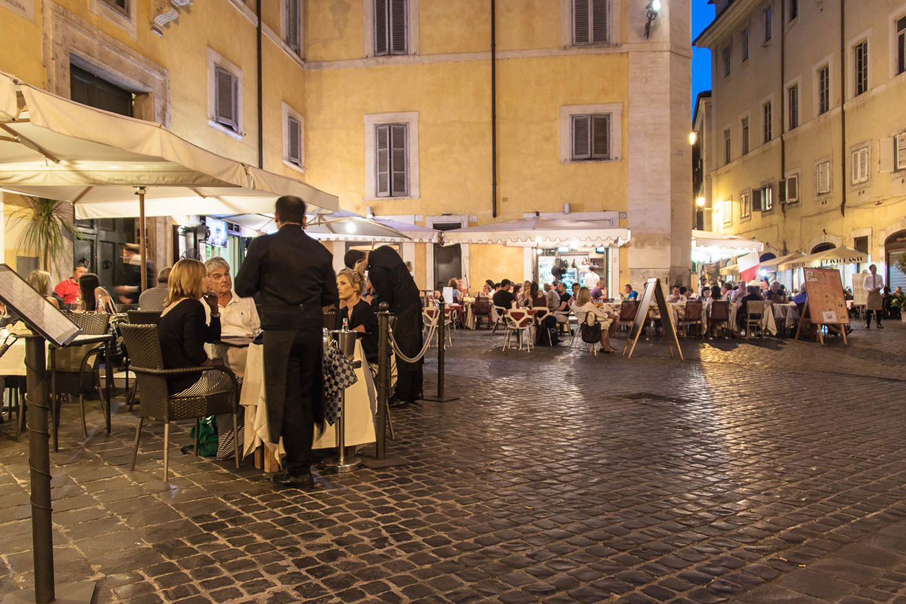 An outdoor dining area in a charming cobblestone square in Rome, bustling with patrons in the evening.