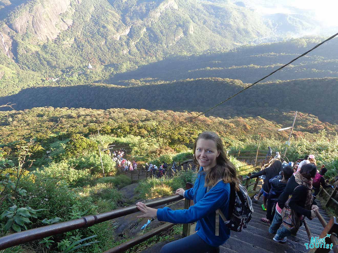 author of the post in a blue jacket and backpack holds a railing while climbing down steps on a mountain trail, with a scenic view of valleys and hills in the background.