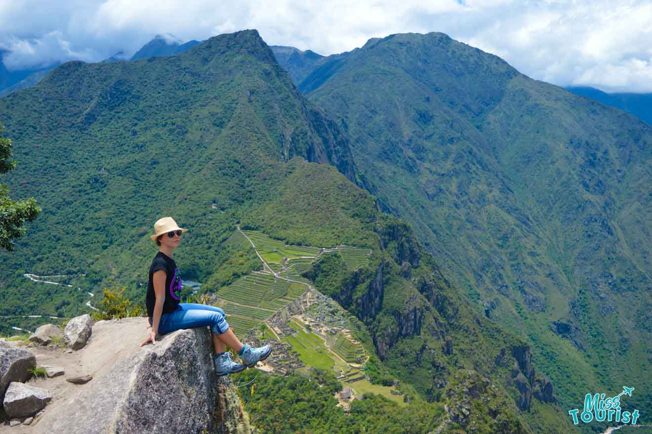 author of the post wearing a hat and sunglasses is sitting at the edge of a cliff overlooking a vast mountainous landscape with visible ancient ruins below.